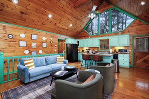 A spacious living area with a blue sofa, a coffee table, and a kitchen with teal cabinets and a large window. - Blue Ridge Retreat - Sauna - Hot Tub - Firepit (Blue Ridge)