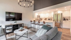 Un salon moderne comprenant un canapé, une table basse et un téléviseur. Il comprend également une cuisine avec des meubles blancs, des appareils en acier inoxydable et un coin bar.