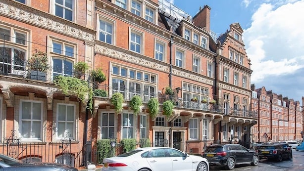 Una hilera de casas adosadas de ladrillo rojo con ventanas de marco blanco, balcones con plantas y coches aparcados en la calle.