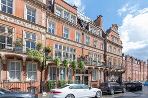 A row of red brick townhouses with white-framed windows, balconies with plants, and parked cars on the street. - Elegant South Kensington Duplex 4-Bed Luxury Stay (London)