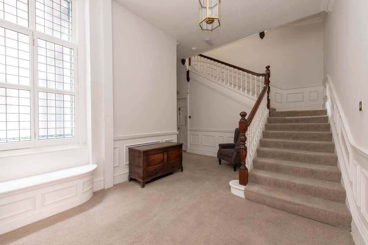 A staircase with a wooden handrail and carpeted steps, a bench, and a wooden cabinet in a room with white walls and a window with a grid pattern.