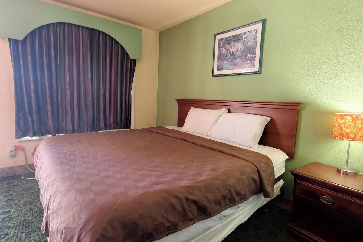 A hotel room with a bed, a nightstand, a lamp, a window with curtains, and a framed picture on the wall.