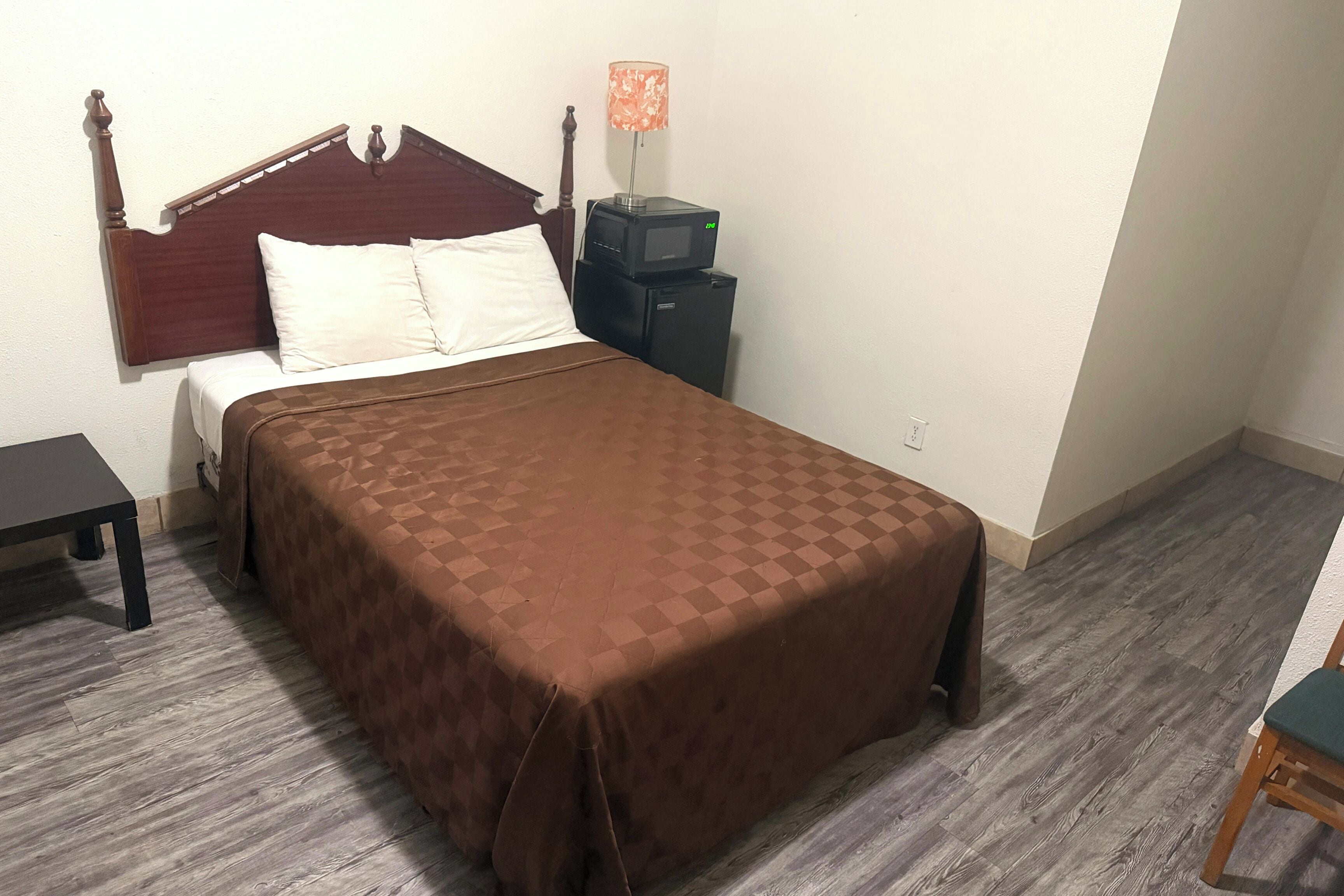 Una cama con cabecera de madera, una mesita de noche, una lámpara y un pequeño electrodoméstico negro en la mesita.