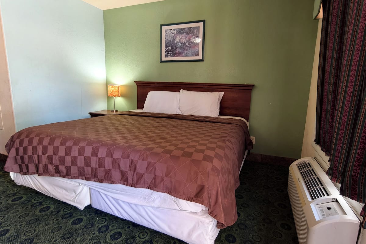 A hotel room with a bed, a nightstand, a lamp, a framed picture on the wall, and an air conditioning unit.