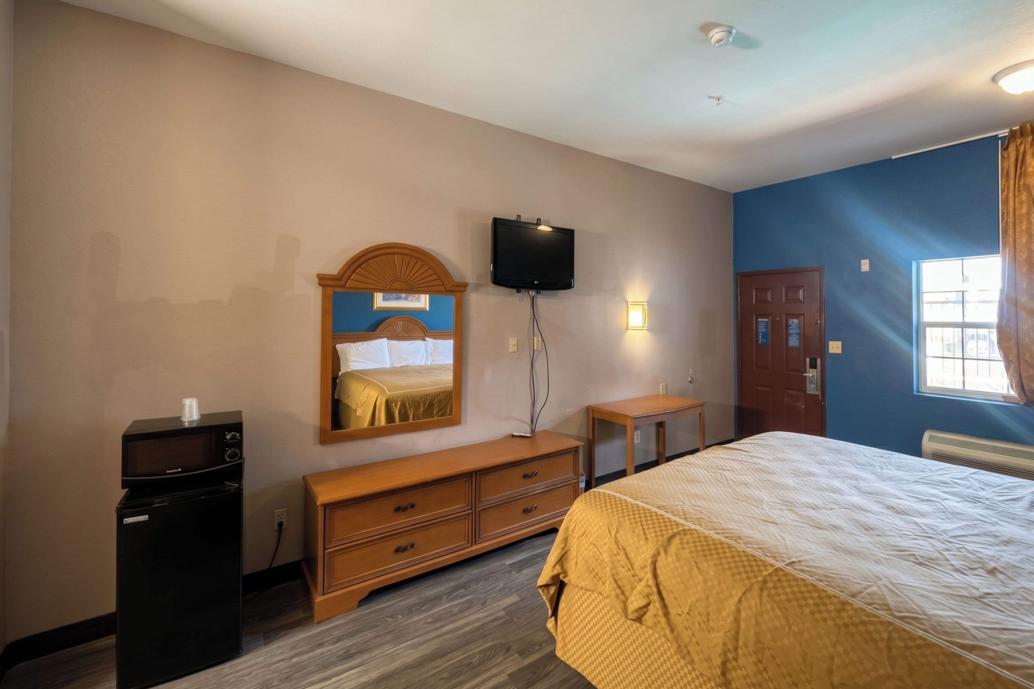 A hotel room with a bed, a television mounted on the wall, a dresser, a desk, and a window with curtains.