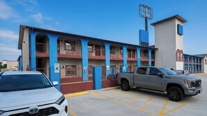 A building with a blue and red facade, a parking lot with marked spaces, and a grey pickup truck parked in one of the spaces. - Airport Inn & Suites South Padre Island I 37 (Corpus Christi)