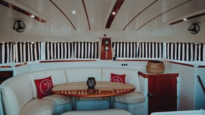 Interior de barco con una mesa redonda, asientos acolchados, un reloj y cortinas a rayas.