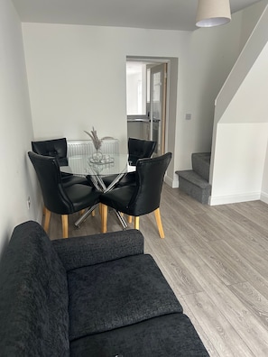 A modern living room with a glass dining table, black leather chairs, a grey sofa, and a staircase leading to an upper level.