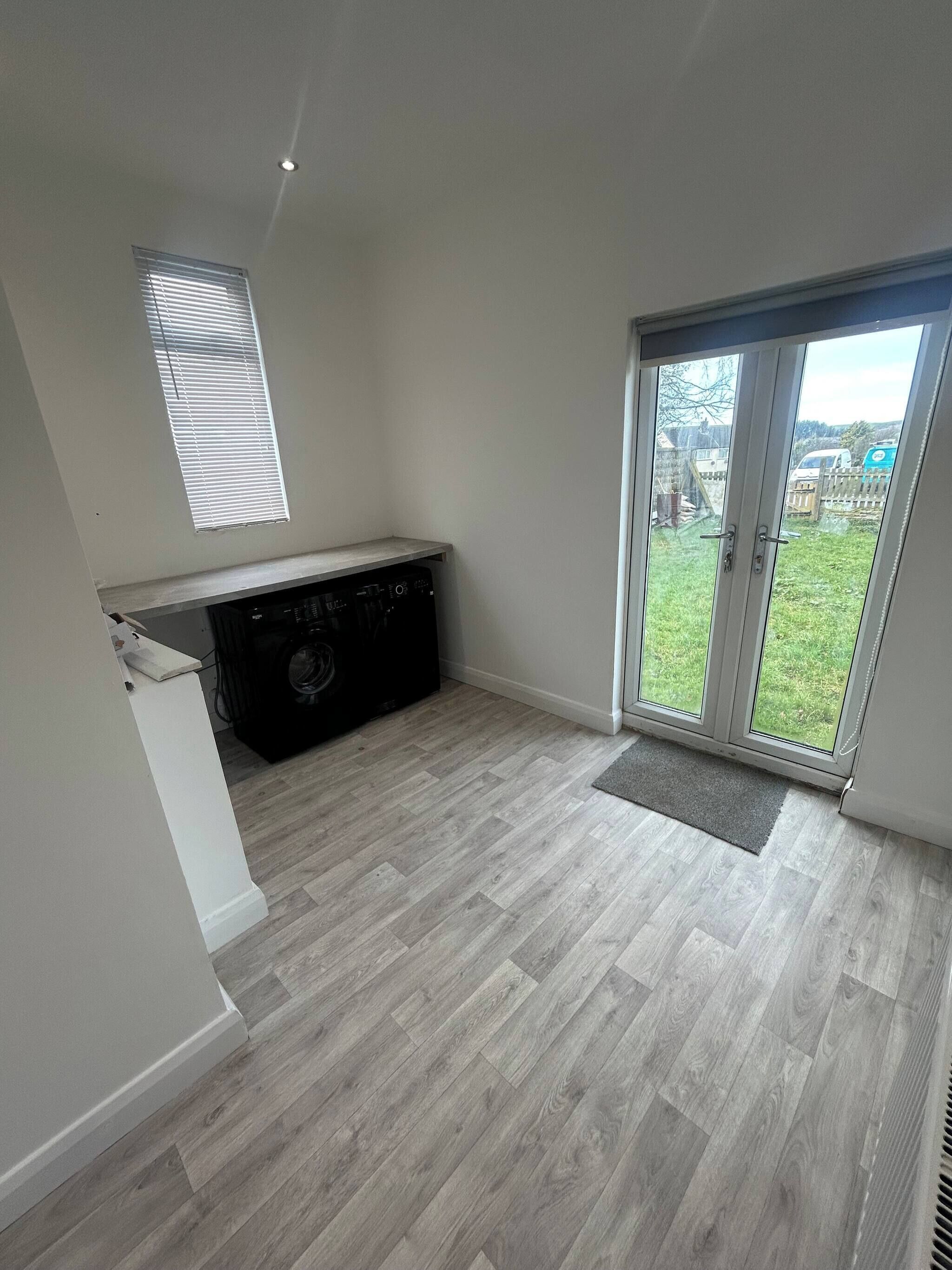 A room with a herringbone-patterned floor, a built-in washing machine, a small window with blinds, and a sliding glass door leading to a backyard.