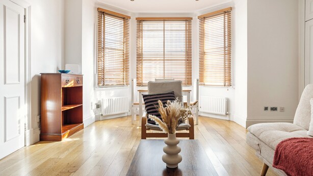 Een kamer met een houten vloer, een witte bank, een stoel met een gestreept kussen, een houten kast en een vaas met gedroogde bloemen op een tafel.