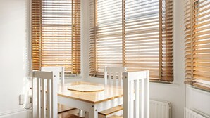 Een eetgedeelte met een houten tafel en stoelen, grote ramen met zonwering en een witte radiator.