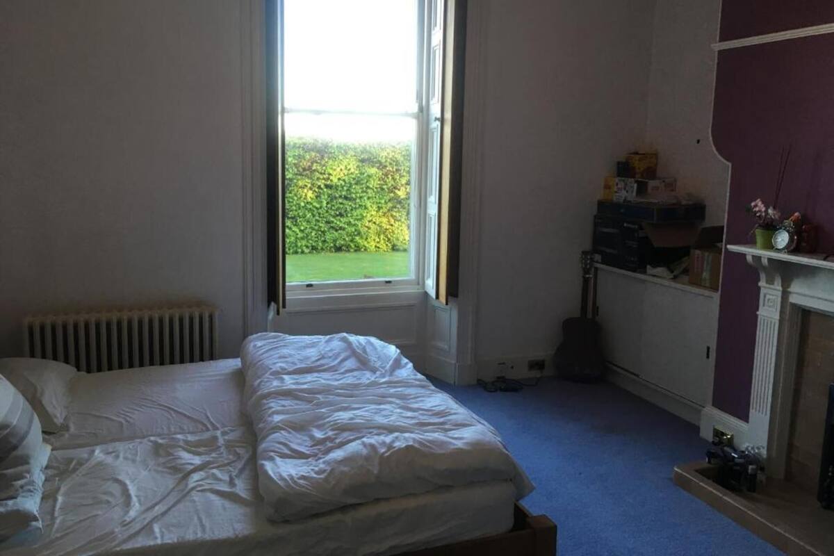 A bedroom with a bed, a radiator, a window with a view of greenery, and a shelf with items.