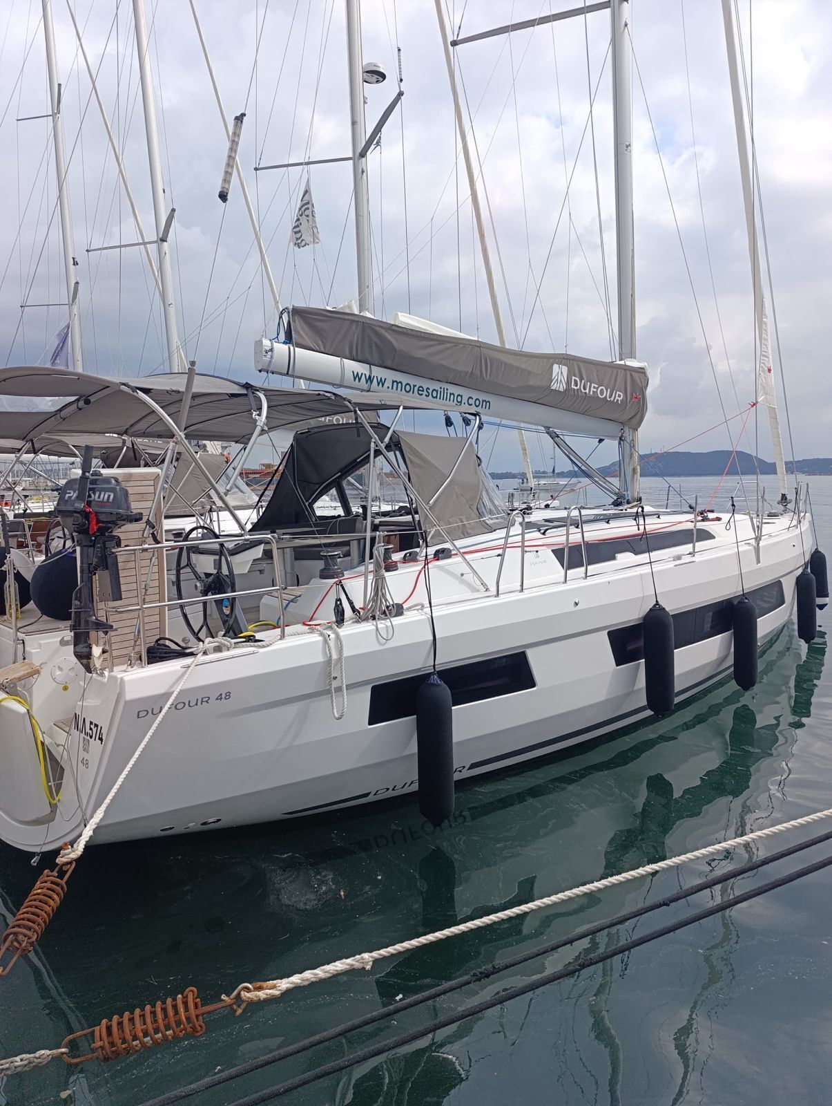 A sailboat docked at a marina with a website banner, a steering wheel, and a winch.