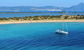 A clear blue sea with a sailboat, a sandy beach, and a distant mountain range.