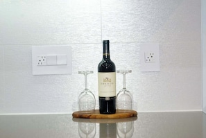A wine bottle on a wooden coaster with two wine glasses on either side, set against a white textured wall with electrical outlets.