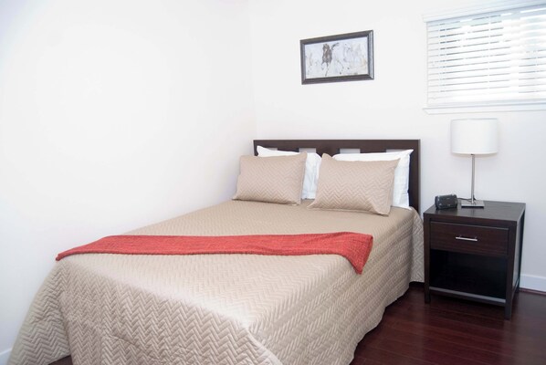 A neatly made bed with a red throw blanket, a bedside table with a lamp, and a framed picture on the wall.