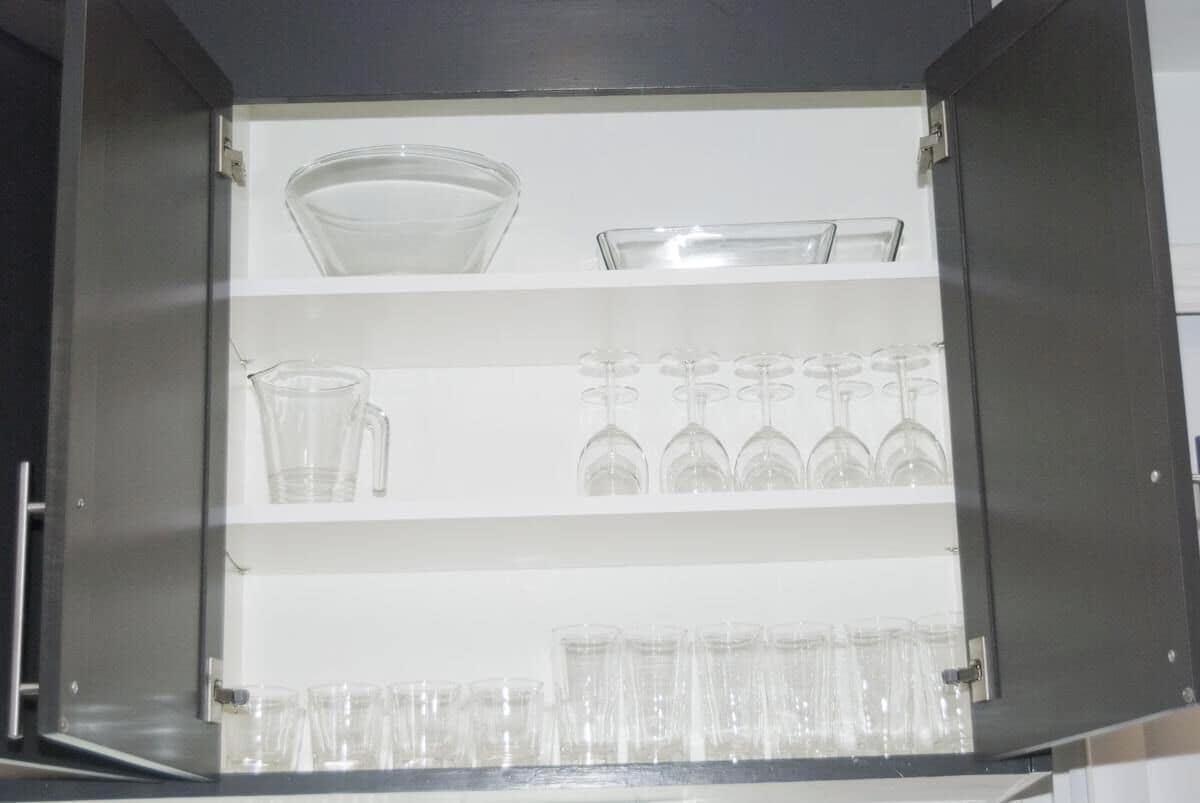 A kitchen cabinet with shelves containing various glassware including a pitcher, wine glasses, and tumblers.