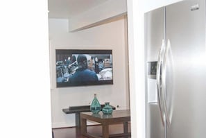 A modern kitchen with a stainless steel refrigerator, a flat-screen TV mounted on the wall, and a wooden table with two teal vases.