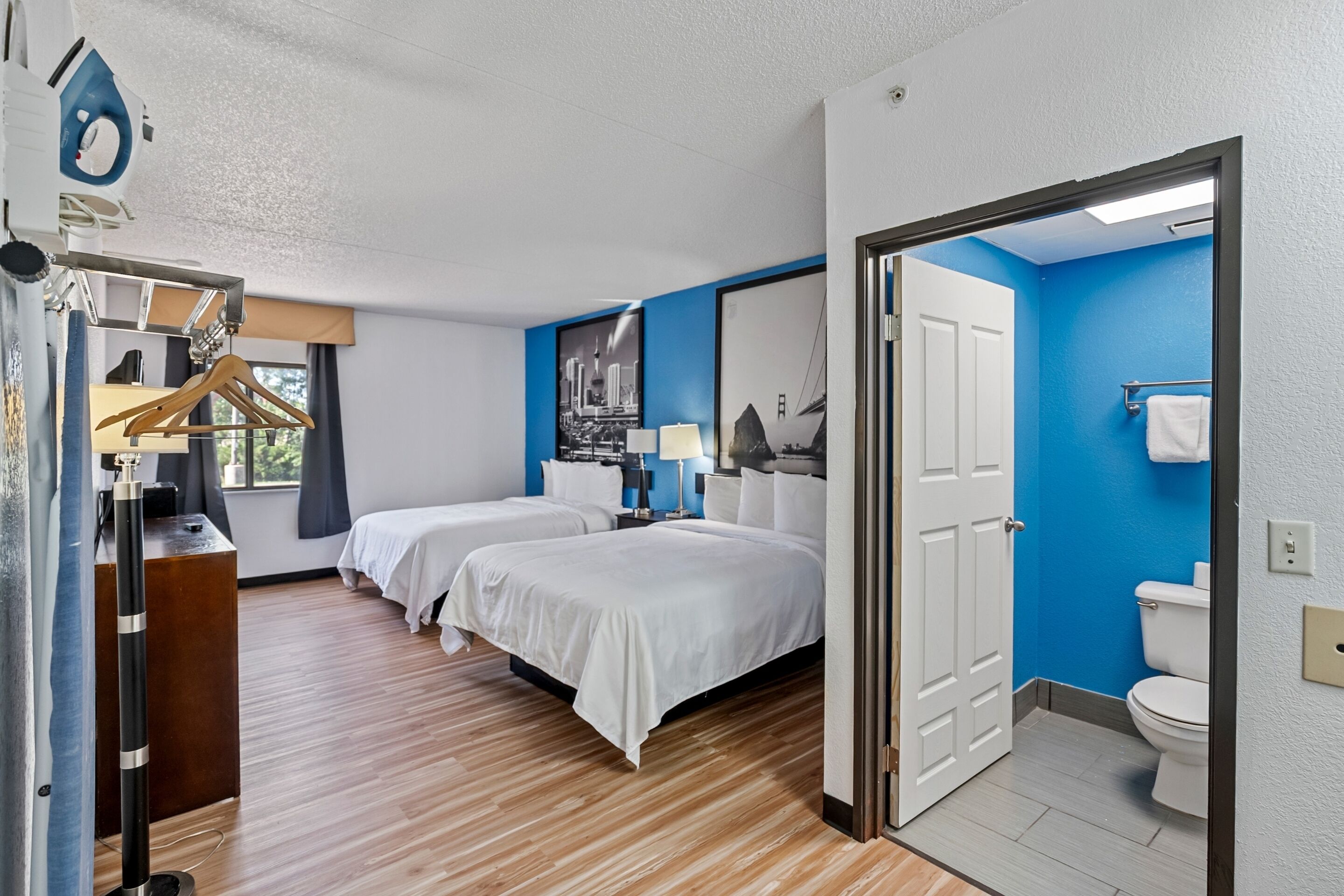 A hotel room with two beds, a desk, a hanging coat rack, and a bathroom with a blue wall and white fixtures.
