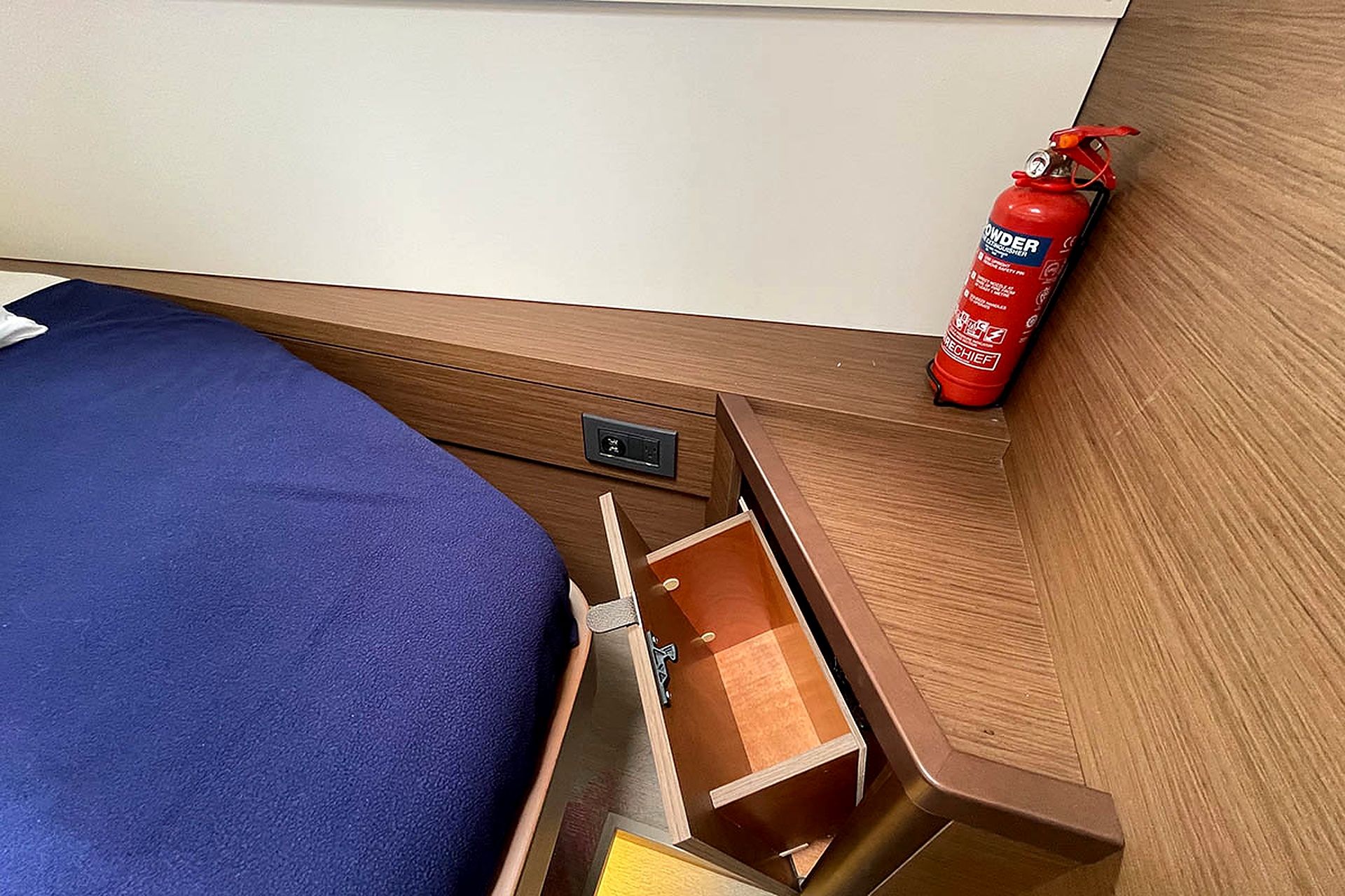 A bed with a blue cover, a wooden nightstand with a drawer, and a red fire extinguisher on the wall.