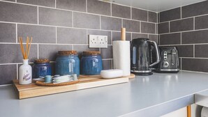 Uma cozinha com azulejos cinzas, uma tomada elétrica branca, tábua de corte de madeira com potes, um rolo de papel toalha e duas chaleiras.
