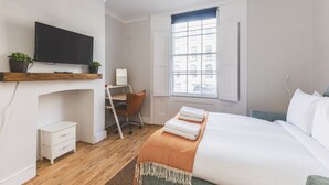 Quarto com cama, escrivaninha, cadeira, TV e janela com vista para edifícios.