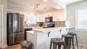 A modern kitchen with stainless steel appliances, a central island with stools, and a view of the adjacent living area. - The Scallop (Carolina Beach)