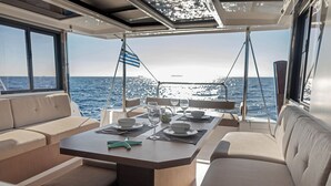 Uma área de refeições em um barco, com uma mesa posta para dois, uma bandeira e vista para o mar.
