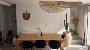 Ein Essbereich mit einem Holztisch, schwarzen Stühlen, einer gewebten Pendelleuchte, einem Kaktus und einer Vase mit Trockenpflanzen.