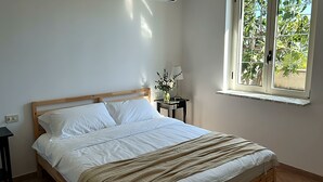 Makuuhuoneessa on puinen sänky, valkoiset vuodevaatteet, yöpöytä, jossa on lamppu ja kukkia, sekä ikkuna, josta avautuu näkymä vehreään maisemaan.