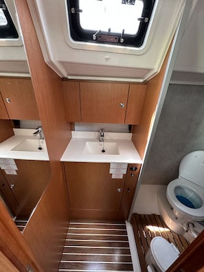 A compact bathroom with a window, two sinks, a toilet, and wooden flooring. - Sun-Kissed Sailing Adventure in Islas Sarónicas (Alimos)