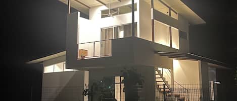 Une maison moderne à deux étages, dotée d’un grand balcon en verre, d’un escalier apparent et d’un jardin clos.