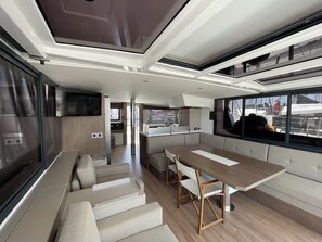A modern interior with a dining area, kitchen, and seating by a large window. - Sun-Kissed Sailing Adventure in Islas Sarónicas (Alimos)
