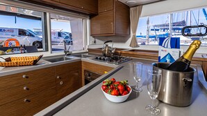 Cucina con pensili in legno, lavello in acciaio inox, piano cottura a gas, una ciotola di fragole, una bottiglia di champagne e una finestra con vista sulle imbarcazioni.
