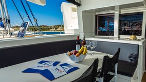 Un tavolo con una ciotola di frutta, bicchieri da vino e una bottiglia, una finestra vista mare e una sedia.