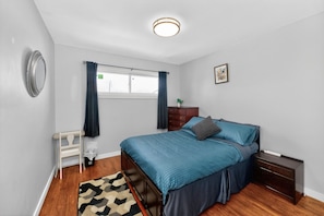 A bedroom with a bed, bedside table, nightstand, window, and a wall-mounted light fixture.