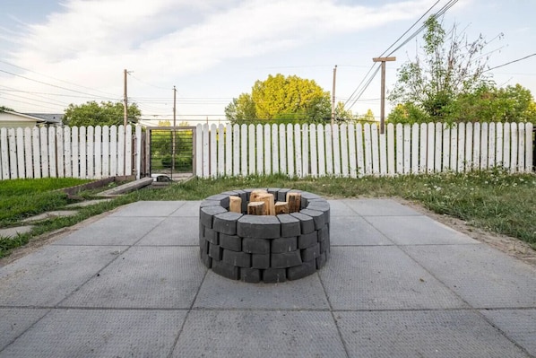 Ein kreisförmiger Feuerkorb aus gestapelten Baumstämmen auf einer Beton Terrasse, umgeben von einem weißen Zaun mit Latten.