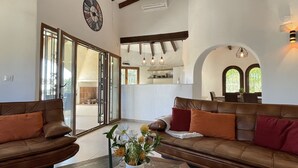 Un'ampia zona giorno con un divano in pelle marrone, un'ampia finestra e una cucina visibile tramite una porta aperta.