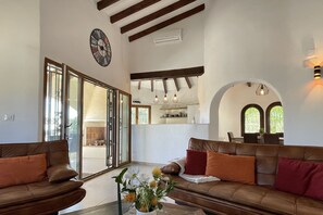 A spacious living area with a brown leather sofa, a large window, and a kitchen visible through an open door. - Private villa with swimming pool for 6 people in Moraira – VILLA ORENSE (Moraira)