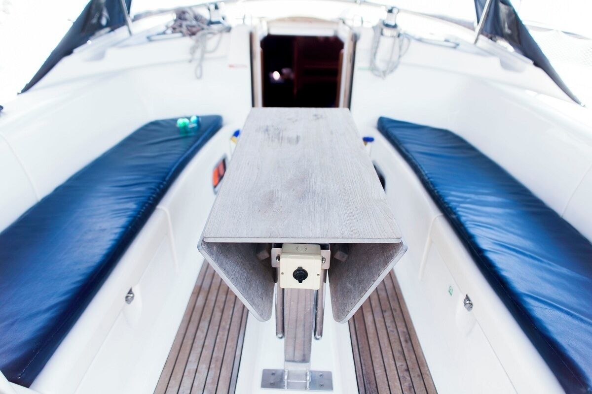 Convés de barco com uma mesa central, duas áreas de assento acolchoadas em azul e entrada para a cabine visível.