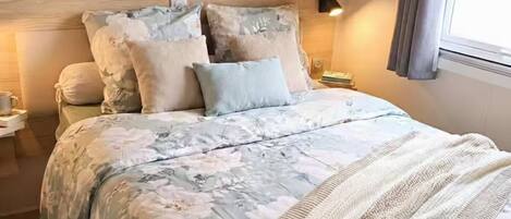 A bedroom with a bed, floral bedding, a knitted throw, wooden headboard, bedside tables, and a window with curtains.