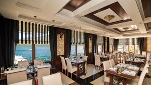 A restaurant with tables set for dining, large windows with curtains, and a view of the sea.