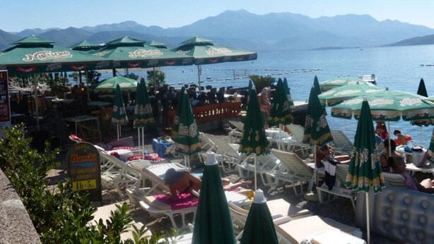 A seaside restaurant with outdoor seating, umbrellas, and a view of the sea and mountains.