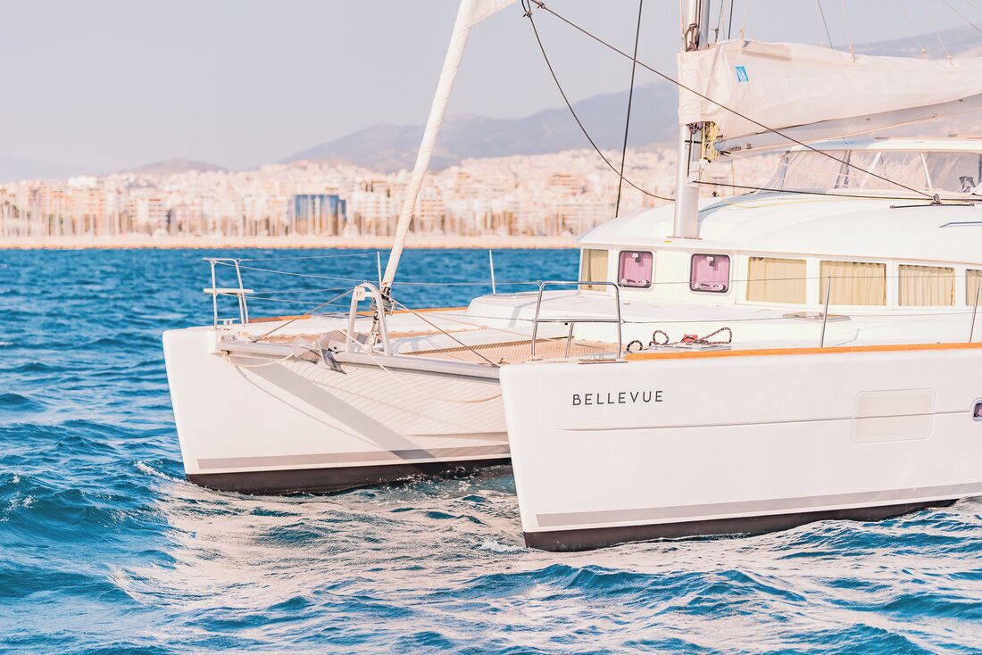 A white sailing boat named 'BELLEVUE' with a deck and cabin structure, sailing on blue water with a cityscape in the background.