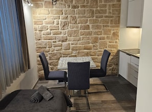 Habitació petita i acollidora amb paret de pedra, taula amb dues cadires, sofà i petita cuina.