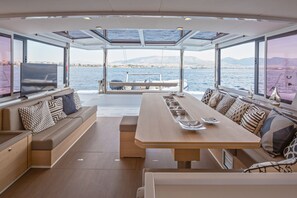 Ein geräumiger Deckbereich mit langem Tisch und Bänken, große Fenster bieten Blick auf das Meer und die Stadt.
