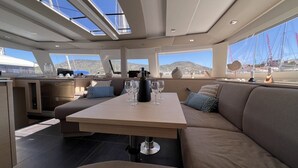 A yacht interior with a dining area, a kitchen, and large windows offering a view of the sea and hills. - Sun-Kissed Sailing Adventure in the Ionian Islands (Nidri)