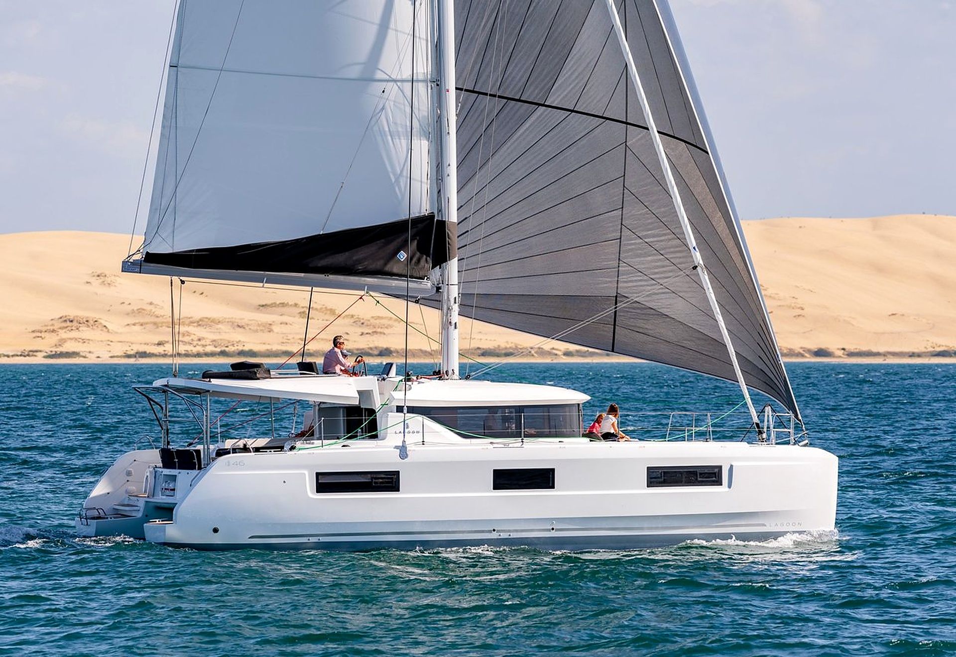 A white catamaran with two visible cabins, sailing on clear blue water with a desert landscape in the background.