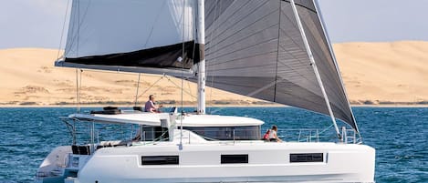 A white catamaran with two visible cabins, sailing on clear blue water with a desert landscape in the background.