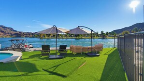 Zona junto a la piscina con césped artificial, sillas de descanso, sombrillas y vistas a un lago y montañas.